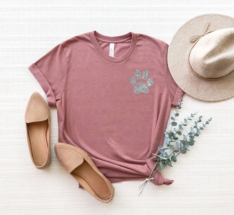Puede incluir: Una camiseta color malva con un dise&ntilde;o de huella de pata de purpurina plateada. La camiseta est&aacute; colocada sobre una superficie blanca con un par de zapatos planos beige y un sombrero de paja.