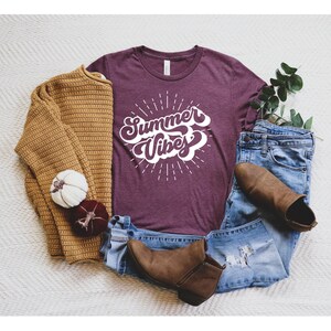 May include: A maroon t-shirt with the text "Summer Vibes" in a white retro font. The shirt is surrounded by a pair of jeans, a brown sweater, and brown ankle boots.