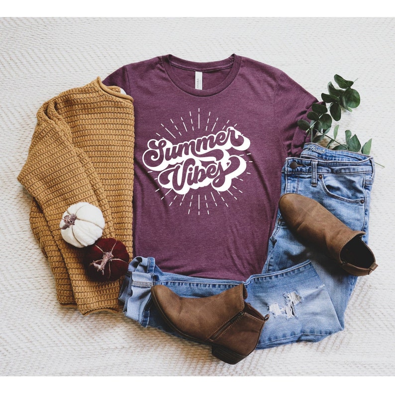 May include: A maroon t-shirt with the text "Summer Vibes" in a white retro font. The shirt is surrounded by a pair of jeans, a brown sweater, and brown ankle boots.