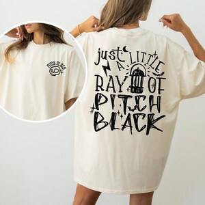 May include: Oversized cream t-shirt featuring black text on the back: "just a little ray of pitch black." The front displays a small skull graphic with "pitch black" text. The shirt is a relaxed fit.