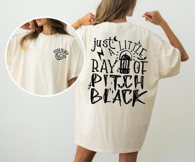 May include: Oversized cream t-shirt featuring black text on the back: "just a little ray of pitch black." The front displays a small skull graphic with "pitch black" text. The shirt is a relaxed fit.