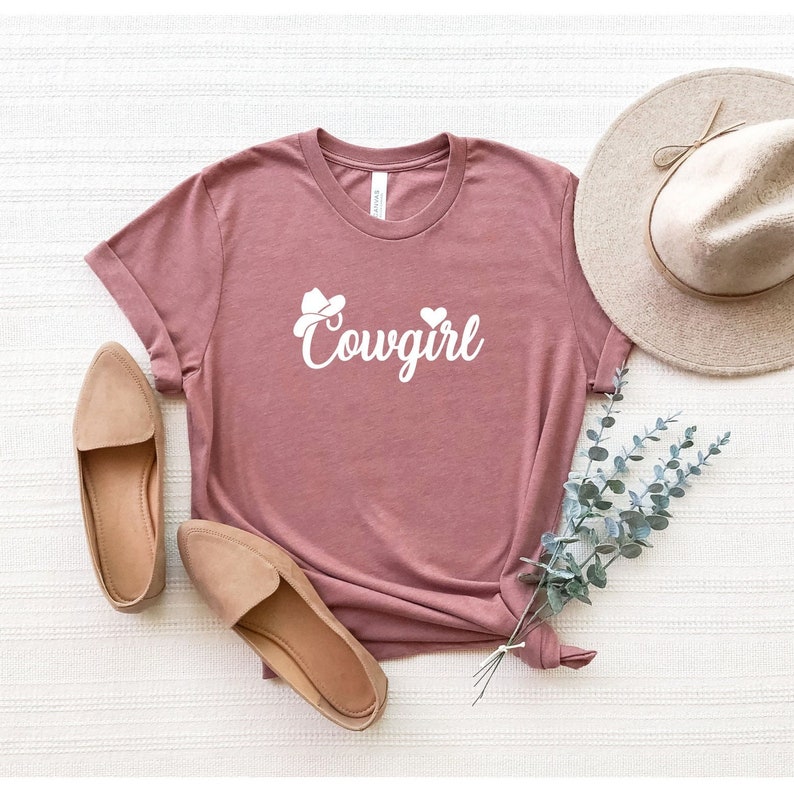 K&ouml;nnte beinhalten: Ein mauvefarbenes T-Shirt mit einer wei&szlig;en Grafik eines Cowboyhuts und dem Wort "Cowgirl" in kursiv.