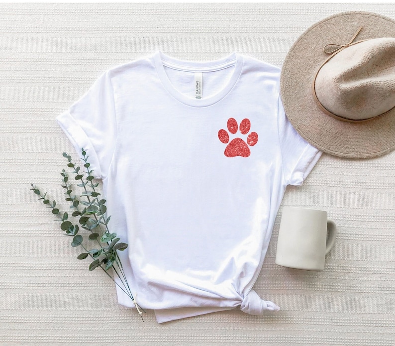Puede incluir: Camiseta blanca con un dise&ntilde;o de huella de pata roja de purpurina.