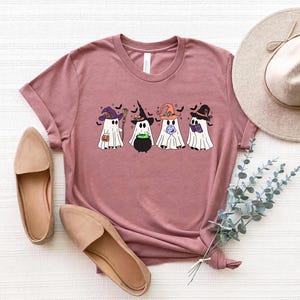 Pu&ograve; includere: Una maglietta color malva con una grafica di cinque fantasmi che indossano cappelli da strega. I fantasmi tengono un calderone, una zucca, una luna e un libro. La grafica &egrave; in stile cartone animato.