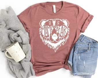 Puede incluir: Una camiseta de color malva con un gráfico de oso blanco. Los ojos del oso están reemplazados por el texto "Mama Bear".