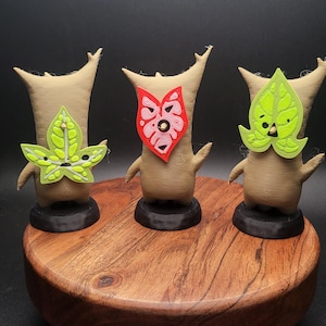 May include: Three small, brown, 3D printed figures with different masks. The figure on the left has a green leaf mask, the middle figure has a pink and red heart mask, and the figure on the right has a green leaf mask.