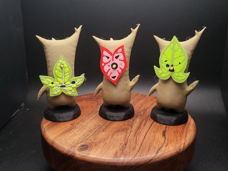 May include: Three small, brown, 3D printed figures with different masks. The figure on the left has a green leaf mask, the middle figure has a pink and red heart mask, and the figure on the right has a green leaf mask.