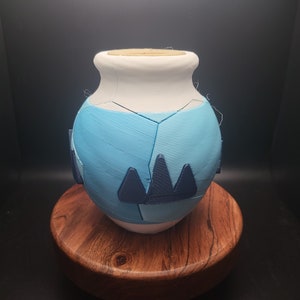 May include: A handcrafted ceramic vase with a white top and a light blue body. The body features geometric designs in a darker blue. The vase is displayed on a wooden stand, showcasing its unique, artistic design.