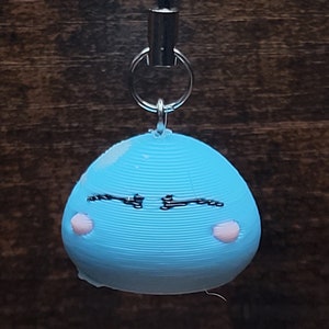 May include: A light blue 3D printed keychain shaped like a round blob with a black drawn face and pink cheeks.