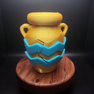 May include: A gold ceramic vase with a blue and white geometric pattern. The vase is sitting on a wooden base.