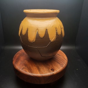 May include: A brown ceramic pot with a yellow glaze, cracked and showing the brown clay underneath. The pot is sitting on a wooden base.