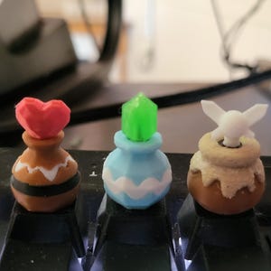 Puede incluir: Tres teclas decorativas para teclado. Una es marrón con un corazón rojo, otra es azul con una gema verde y la última es marrón con un hada blanca. Están diseñadas para encajar en un teclado mecánico.
