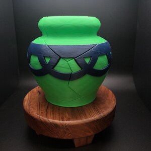 May include: A green ceramic pot with a dark blue design around the middle. The pot is sitting on a wooden base.