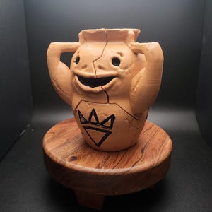May include: A brown ceramic figurine of a smiling character with arms raised. The character has a crown and a triangle symbol on its chest. The figurine is sitting on a wooden base.