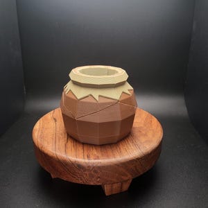 Puede incluir: Un pequeño macetero geométrico de color terracota con un borde marrón claro se asienta sobre un soporte de madera. El macetero tiene un diseño único y facetado.