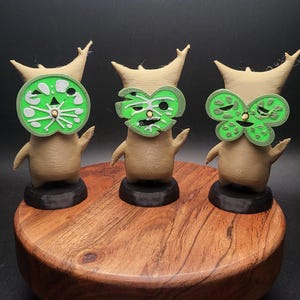 May include: Three small, brown, stylized figures with green masks. The masks are different designs, one is a circle, one is a heart, and one is a butterfly. The figures are standing on a wooden base.