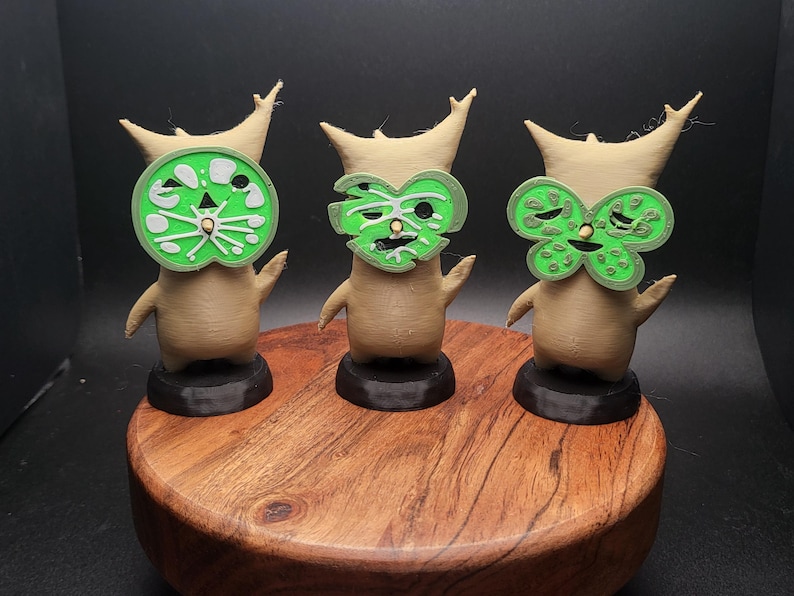 May include: Three small, brown, stylized figures with green masks. The masks are different designs, one is a circle, one is a heart, and one is a butterfly. The figures are standing on a wooden base.