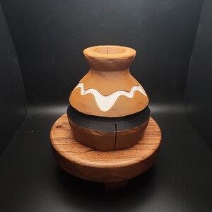 May include: A small, brown ceramic pot with a white and black design sits on a wooden base. The pot has a rounded shape and a wide opening at the top.