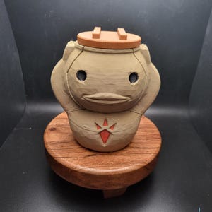 Könnte beinhalten: Eine handbemalte Holzfigur eines Videospielcharakters. Die Figur ist in Braun- und Beigetönen bemalt, mit einem roten Diamanten auf der Brust. Die Figur steht auf einem Holzsockel.