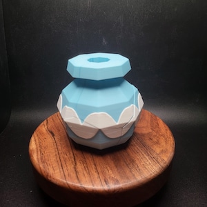 May include: A light blue and white geometric vase with a decorative scalloped edge. The vase is displayed on a round, wooden stand with a dark background. The vase has a unique, polygonal design.