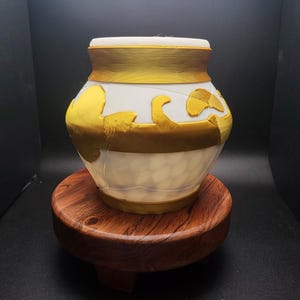 May include: A white ceramic jar with gold accents and a decorative design. The jar is sitting on a wooden base.