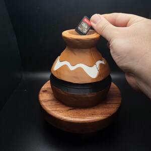 May include: A brown and black ceramic pot with a white design, sitting on a wooden base. A video game cartridge is being placed into the pot.