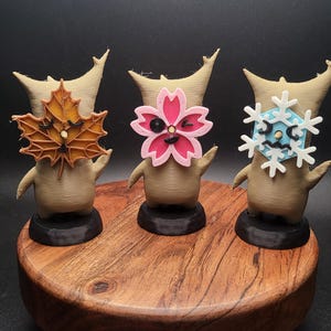 May include: Three small, brown, stylized figures with different colored faces. The figure on the left has a brown leaf face, the middle figure has a pink flower face, and the figure on the right has a blue snowflake face. The figures are standing on a wooden base.