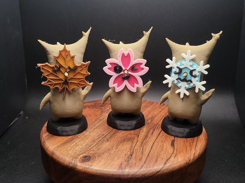 May include: Three small, brown, stylized figures with different colored faces. The figure on the left has a brown leaf face, the middle figure has a pink flower face, and the figure on the right has a blue snowflake face. The figures are standing on a wooden base.