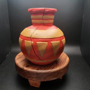 May include: A red and gold ceramic vase with a geometric pattern sits on a wooden stand. The vase is cracked down the side.