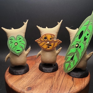 May include: Three small, beige, stylized figures with green and brown leaf-shaped masks. The figures are standing on black bases and are arranged on a wooden surface.