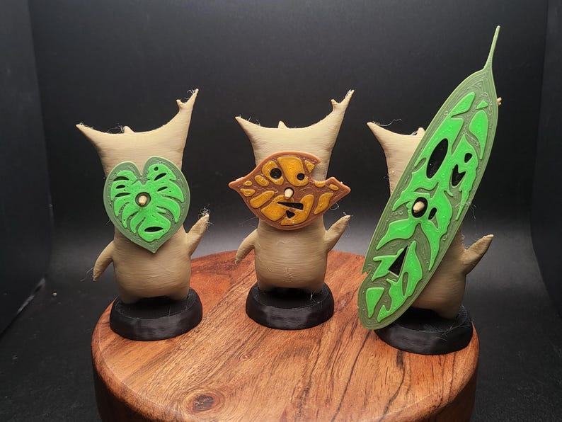 May include: Three small, beige, stylized figures with green and brown leaf-shaped masks. The figures are standing on black bases and are arranged on a wooden surface.