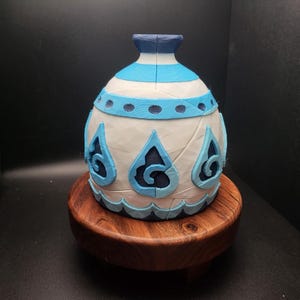 May include: A white ceramic vase with blue and white geometric designs. The vase has a round body and a narrow neck. It is sitting on a wooden base.