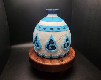May include: A white ceramic vase with blue and white geometric designs. The vase has a round body and a narrow neck. It is sitting on a wooden base.