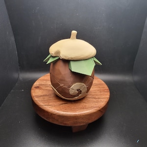 May include: A brown and white ceramic acorn-shaped container with a green leaf lid, sitting on a wooden pedestal.