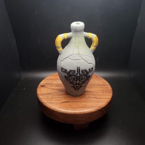 May include: A white ceramic bottle with gold handles and a black design. The bottle is sitting on a wooden base.