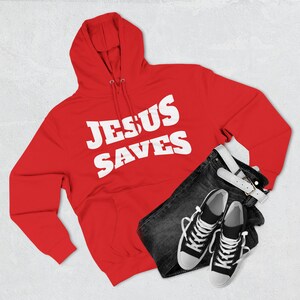 Könnte beinhalten: Ein roter Kapuzenpullover mit dem weißen Text "JESUS SAVES". Das Bild zeigt auch schwarze Jeans, einen weißen Gürtel und schwarz-weiße Turnschuhe. Die Artikel sind auf einer hellgrauen Oberfläche angeordnet.