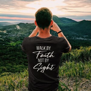 May include: A person wearing a black t-shirt with the text "Walk by Faith not by Sight" printed on the back. The person is standing on a hilltop overlooking a valley with a sunset in the background.