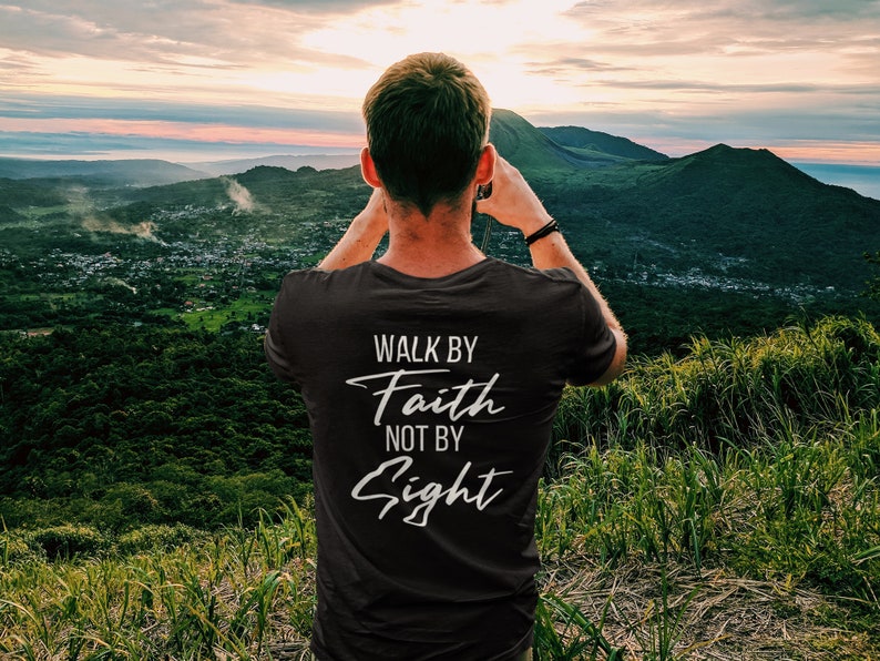 May include: A person wearing a black t-shirt with the text "Walk by Faith not by Sight" printed on the back. The person is standing on a hilltop overlooking a valley with a sunset in the background.