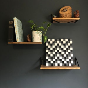 May include: Two wooden shelves with black metal brackets mounted on a dark grey wall. The top shelf has a wooden sculpture of a snake, a small wooden bird figurine, and a small metal object. The bottom shelf has a black and white chequered wall art piece and a potted plant with green leaves.