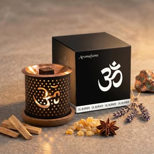 May include: Aroma diffuser with a black and copper design. The diffuser has a cylindrical shape with a perforated pattern and the Om symbol. A black box with the word "Aromafume" and the Om symbol is behind the diffuser. The words "OIL BURNER" are printed on the box.