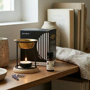 May include: An essential oil burner with a gold-colored bowl and a black metal frame. A lit candle sits below the bowl. A bottle of essential oil and a black box with the text "Aromafume" are also present. The burner is on a wooden surface.