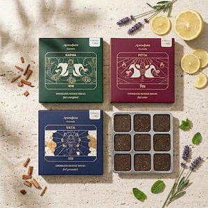 May include: An overhead shot of four boxes of Aromafume Ayurveda smokeless incense bricks. The boxes are green, burgundy, and navy blue, with gold accents. A metal tray of incense bricks is in the center. The image includes lemons, lavender, and mint.