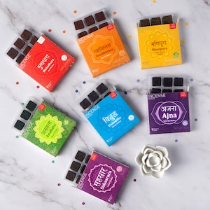 May include: A set of nine incense bricks in individual boxes, each with a different color and design. The boxes feature the names of the seven chakras in Sanskrit, along with their corresponding English names and chakra attributes. The boxes are arranged in a circle on a white marble surface.