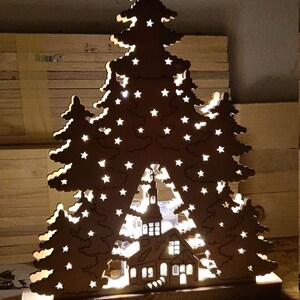 May include: A wooden Christmas tree silhouette with a light-up house scene in the center. The tree is decorated with stars and has a rustic brown finish.