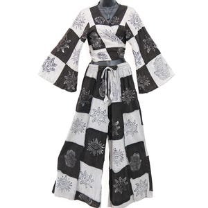 Op de afbeelding: Een tweedelige outfit bestaande uit een wikkeltop met lange mouwen en een broek met wijde pijpen. Beide kledingstukken hebben een zwart-wit geblokt patroon met prints van zonnen, ogen en hamsa-handen. De broek heeft een elastische tailleband.