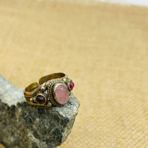 May include: A silver ring with a pink oval gemstone and two smaller red gemstones. The ring is on a gray rock.
