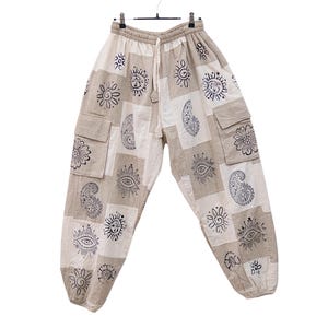 May include: Beige and off-white patchwork cargo pants featuring a repeating black ink print of suns, eyes, and paisley patterns. The pants have an elastic waistband with a drawstring, side cargo pockets, and elasticized cuffs at the ankles.