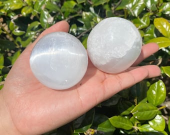 Op de afbeelding: Twee witte seleniet bollen, een met een glad oppervlak en de andere met een gestreept oppervlak, worden getoond op een hand tegen een groene, bladerige achtergrond.