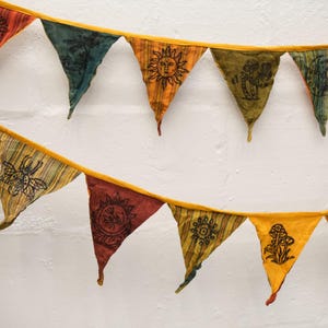 May include: A fabric bunting banner featuring two rows of triangular flags in shades of orange, yellow, green, and red. Each flag displays a black ink-style print, including images of a sun, mushrooms, a beetle, and various celestial symbols.