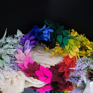 May include: A rainbow-colored felt flower garland with leaves. The garland is made of felt flowers in shades of blue, purple, pink, red, orange, yellow, and green.